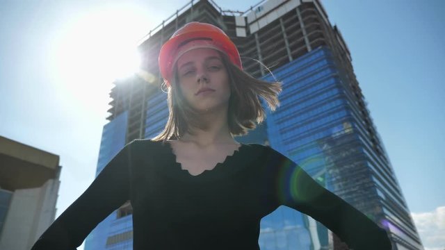 Young confident girl builder with hemlet is standing with hands on hips in daytime in summer, turning head, building concept, urban concept, building on background