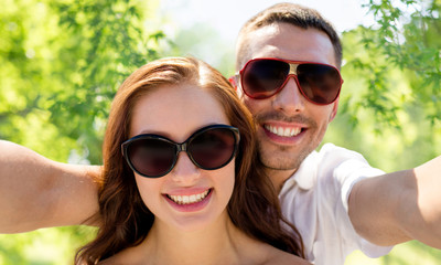 love, summer and people concept - smiling couple wearing sunglasses making selfie over green...