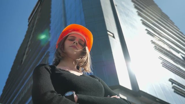 Young girl builder with hemlet is standing with arms crossed in daytime in summer, building concept, urban concept, building on background, bottom view