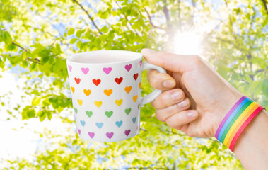 homosexual and lgbt concept - female hand holding cup with rainbow colored heart pattern and gay pride awareness wristband over natural summer background