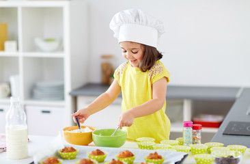family, cooking, baking and people concept - little girl in chefs toque making batter for muffins...