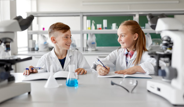 education, science and children concept - happy smiling kids with workbooks studying chemistry at school laboratory and talking