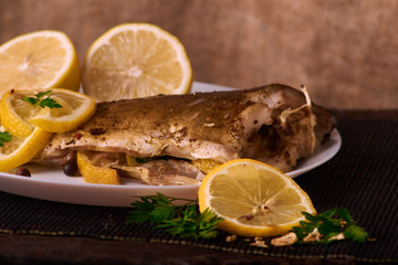 Baked fish with lemon, spices and herbs on white plate on wooden background without head