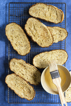 Chargrilled oblong bruschetta bases on a wire rack with a bowl of oil and a pastry brush.