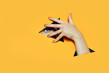 A girl with beautiful long fingers and gold nail varnish covers her eyes in a triangular hole of orange paper.