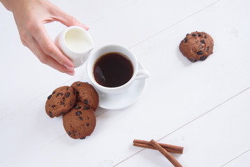 A woman's hand pours milk into coffee. A cup of fragrant coffee with cinnamon and cookies on a white background. Top view.