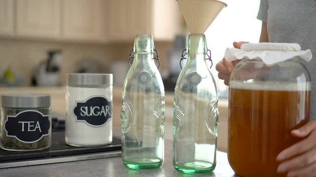 Female brings a large jar of kombucha tea and pours it into two glass bottles 