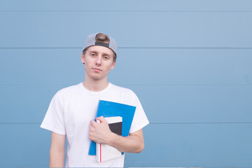 Serious young man in a white T-shirt stands on a blue background with books and notebooks in his hands and looks at the camera.Portrait of a student with books against the background of a blue wall.