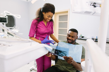 Obraz premium Young African woman dentist in a pink suit and blue medical gloves preparing for a patient treatment and speaking with african young man patient