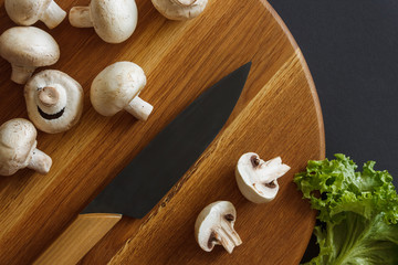 Still life of vegetables. A wooden board, which contains champignons, cabbage leaves and a kitchen knife. Dark background