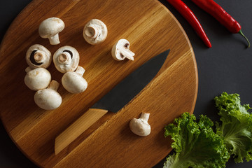 Obraz na płótnie Canvas Still life of vegetables. A wooden board, which contains champignons, cabbage leaves, pods of acute red pepper and a kitchen knife. Dark background