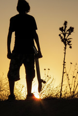 young skater holding his skateboard in his hand against the light watching a beautiful sunset next to a plant with spikes