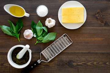 Ingredients for pesto sauce. Cheese, garlic, green basil, olive oil, salt near grater and mortar on dark wooden background top view copy space