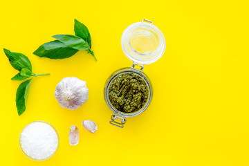 Composition with green pesto sauce in glass jar, basil leaves and garlic on yellow background top view copy space