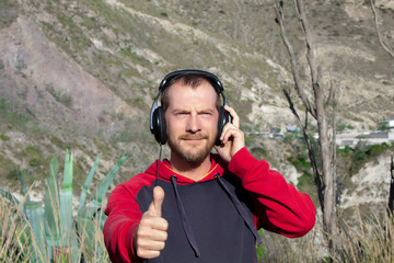 A bearded man listens to music on headphones, in nature. There are mountains behind it. He shows his thumb up.