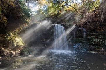 Small waterfall from a thermal spring with early morning sunbeams streaming through the trees and illuminating the steam.