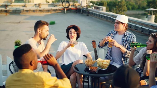 Happy men and women are clinking glasses and bottles with drinks then drinking and talking sitting on roof at table on sunny day. Party, beverage and conversation concept.