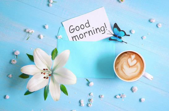 Cup of cappuccino coffee, lily flower and  inscription "Good morning" on blue wooden background. Concept of morning, top view, flat lay.