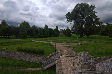 Fototapeta na wymiar Ancient road through the park to the Church of Exaltation of the Holy and St. Joseph. View from terrace of Pidhirtsi Castle. Heavy clouds in the sky. Several minutes before rain. Pidhirtsi, Ukraine