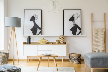 Real photo of a small table standing between a pouf and baskets on a white rug and in front of a cupboard and shelf with posters standing between a lamp and a ladder in living room interior