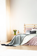 Grey blanket on pink bed with headboard in pastel bedroom interior with flowers on table. Real photo