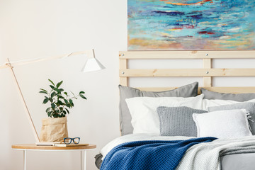 Plant and lamp on table next to bed with blue blanket in bedroom interior with painting. Real photo