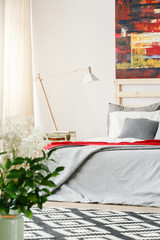Lamp next to grey bed in bright bedroom interior with red painting and flowers. Real photo