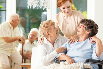 Happy friendship in old age. Tender caregiver standing behind senior women hugging each other in a...