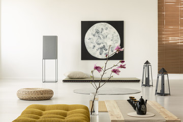 Kettle on wooden desk next to yellow pouf and flowers in japanese interior with moon poster. Real photo