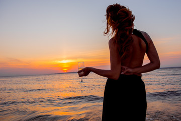 woman at sunset. Elegant girl is drinking champagne at sunset. Girl with  glass on background setting sun. romantic date in the evening. Luxury woman drinking from a wine glass on the beach. 
