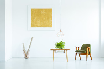 Real photo of a green armchair standing next to a table with a plant in white living room interior with a mockup poster