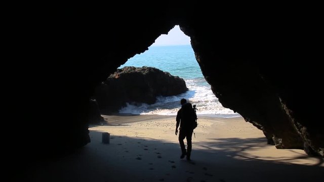 A hiker walks through a large cave and out onto a beach by the ocean.
