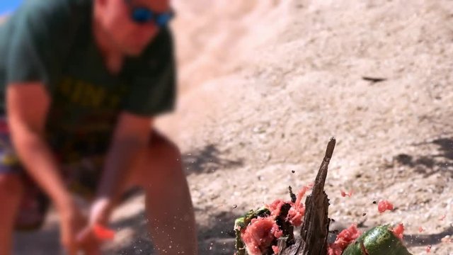Slow motion: Sexy young guy in summer clothes and glasses, destroys watermelon with a baseball bat. It is located on a sandy beach near the sea in summer.