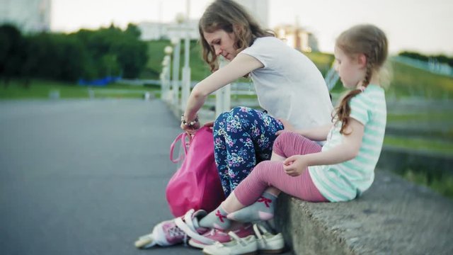 Mother helps his daughter to wear a helmet and protective gear, for roller skating in the park. Woman helps girl put on protective knee and elbow pads. Active family rest in the park.