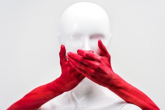 cropped image of woman in red paint covering mannequin mouth with hands isolated on white