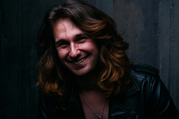 brutal man in a leather jacket on a naked body with long hair. a lonely man is sad and desperate. smiles, leans against the wall