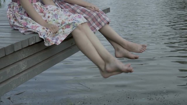 Two children shake their bare feet above the water. They sit on a wooden bridge over the water next to each other. Close up. Raw video. 4K. 29.97 fps.