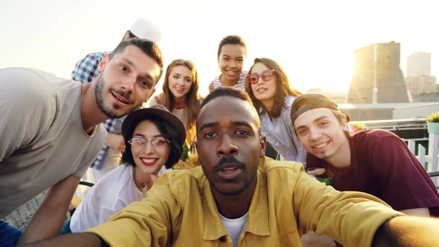 Point of view shot of handsome African American man taking selfie with his happy friends, people are looking at camera, smiling and posing with bottles at roof party.