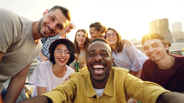 Point of view shot of young people multiethnic group taking selfie and holding camera, men and women are looking at camera, smiling and posing with drinks at rooftop party.