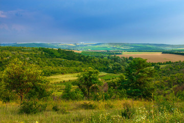 Fototapeta premium summer landscape. The dark sky is covered with storm clouds and the bright green and yellow fields lie in the hills
