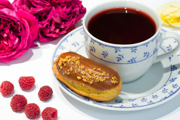 A cup of tea with caramel eclair, raspberry and chocolate. Beautiful desing with pink and yellow roses.