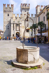Fototapeta na wymiar The town's water well and market square with in the background the front gate, draw bridge and bastions of the walled town of Sirmione, Lombardy, Italy