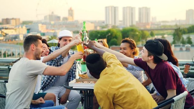 Young people are toasting, clinking bottles and drinking beer and sodas, eating snacks, chatting and laughing during rooftop party in summer.