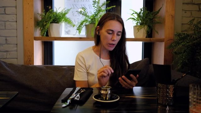 Young woman sitting in cafe and learning language using an app on her smartphone. She is listening to new words in headphones and then repeats them while drinking coffee