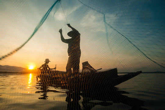Asian fisherman on wooden boat casting a net for catching freshwater fish in nature river in the early morning before sunrise.Fishermen fishing in the morning light.