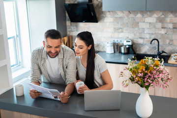 portrait of smiling married couple reading newspaper together at counter with laptop in kitchen