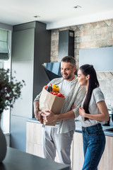 happy married couple with paper package full of fresh vegetables for dinner in kitchen at home