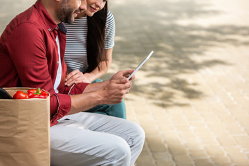 partial view of married couple using tablet together while resting after shopping on bench on street