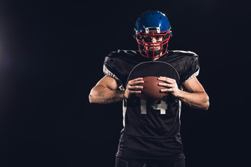 confident american football player holding ball in hands and looking at camera isolated on black