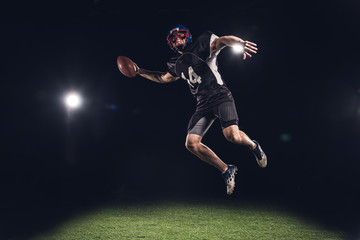 Fototapeta na wymiar american football player jumping with ball under spotlights on black